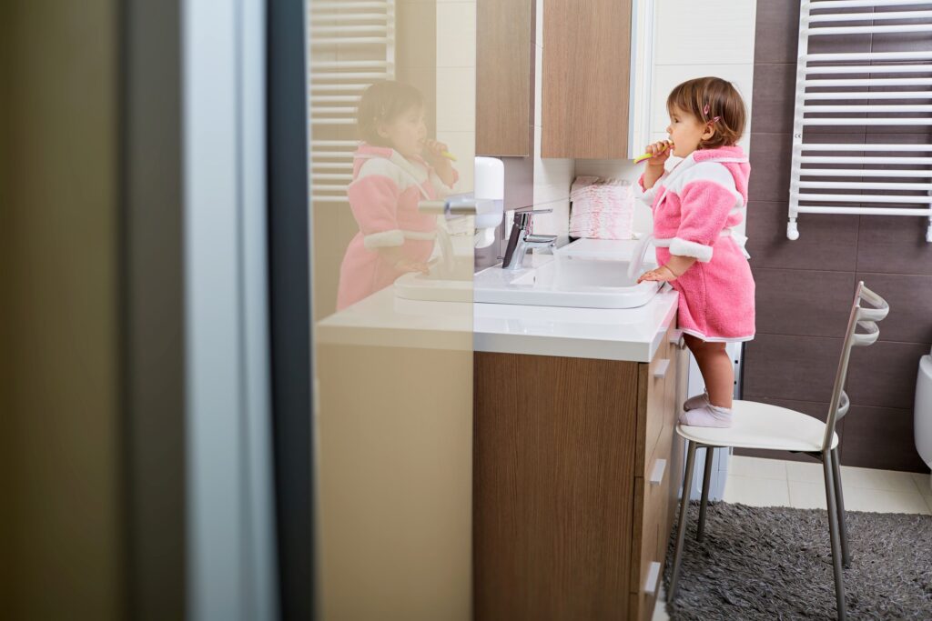 Little girl in pink robe on chair brushing teeth looking at her reflection